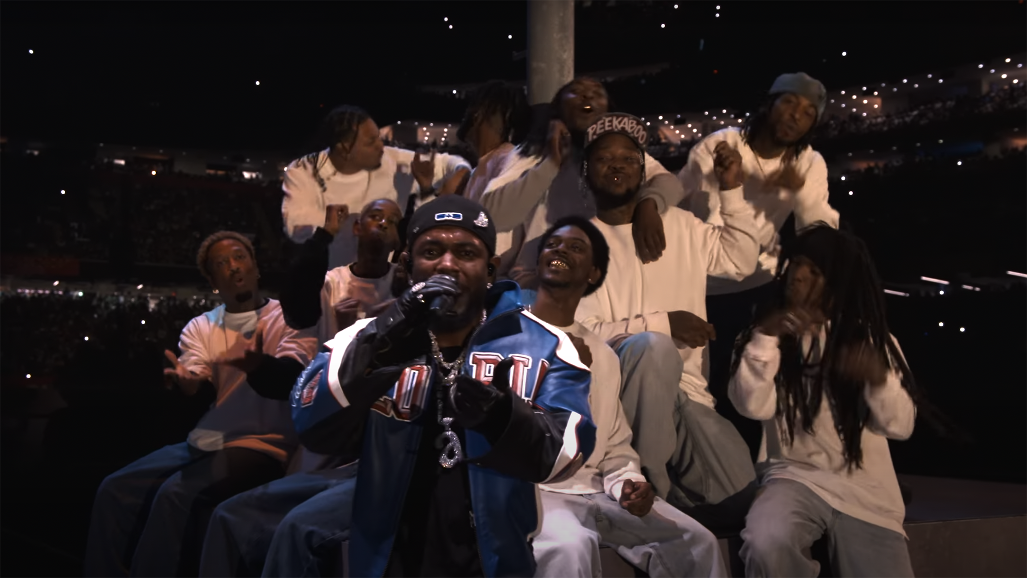 A group of dancers in white T-shirts and gold teeth pose behind Kendrick Lamar as he raps.
